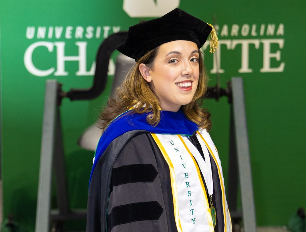 Kaitlin Klotz wearing her doctoral regalia with graduate stoles, posing in front of the UNC Charlotte victory bell 