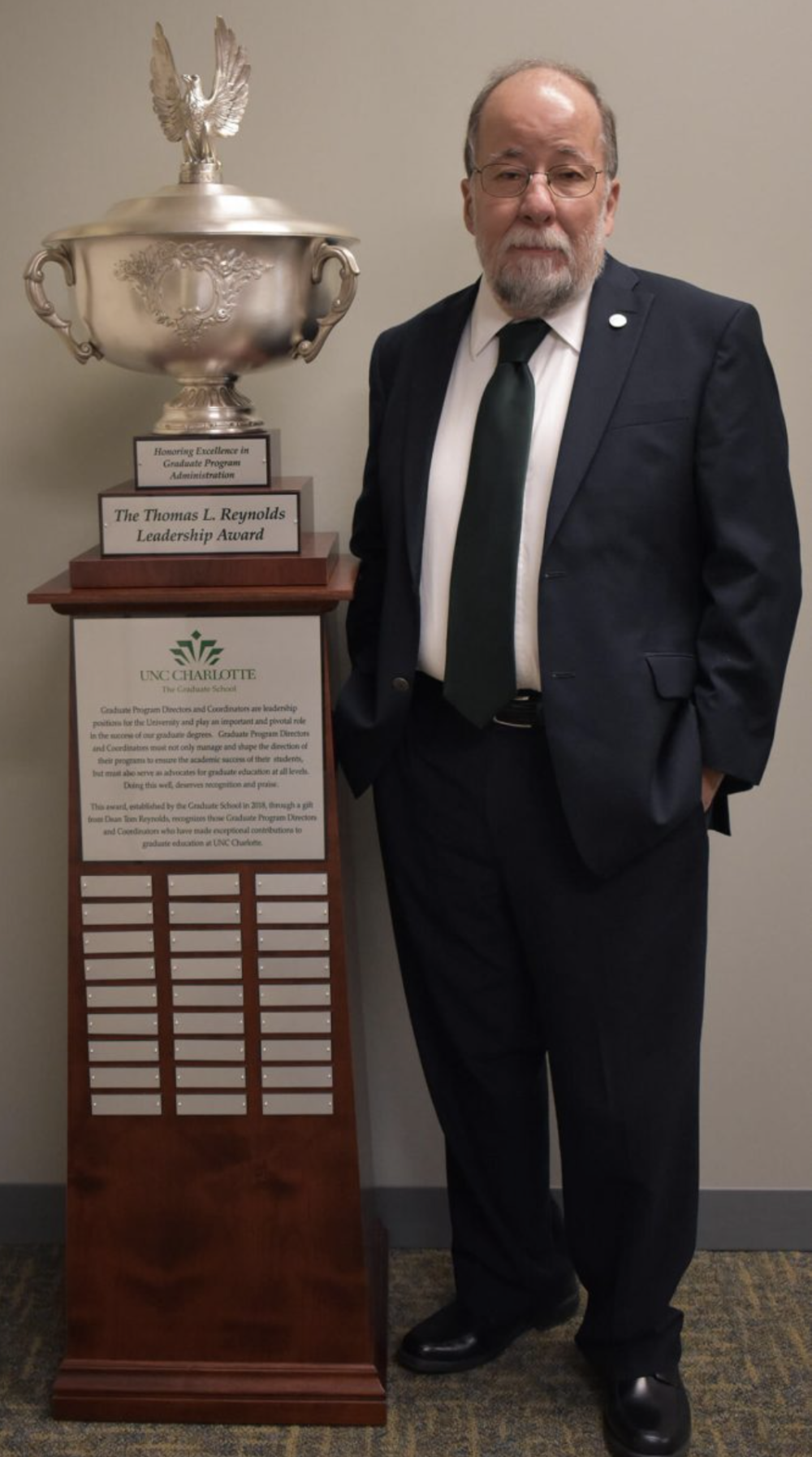 Dean Emeritus Tom Reynolds poses with hands in pockets and trophy to the left of the image