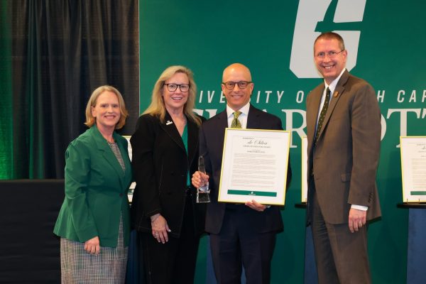Four university officials standing on a stage posing for a photo as a man in the center receives a framed Graduate Mentor Award certificate and a glass trophy.